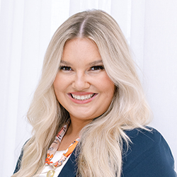 A woman with long blonde hair wearing a navy blazer and patterned scarf, smiling in front of a white curtain background.