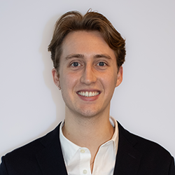 A young man with light brown hair, wearing a dark blazer and white shirt, smiles at the camera against a plain light background.