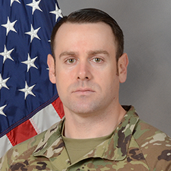 A man in military uniform stands in front of a U.S. flag, facing the camera with a neutral expression.