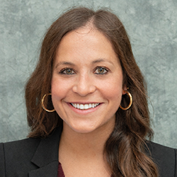 A woman with long brown hair, wearing a black blazer and gold hoop earrings, smiles in front of a mottled gray background.