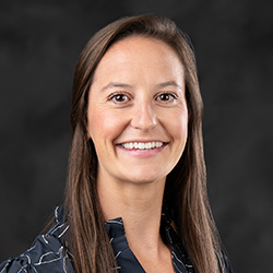 A woman with long brown hair smiles at the camera. She is wearing a dark blouse with a light pattern and is posed against a dark, plain background.