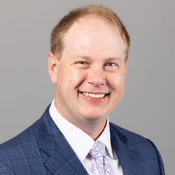 A man with short light hair wearing a blue suit, white shirt, and patterned tie, smiling at the camera against a plain gray background.
