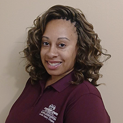 Woman with curly hair smiles at the camera, wearing a maroon collared shirt with a white logo, standing against a plain beige wall.