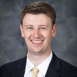 A man in a dark suit, white shirt, and light yellow tie smiles at the camera against a gray studio background.