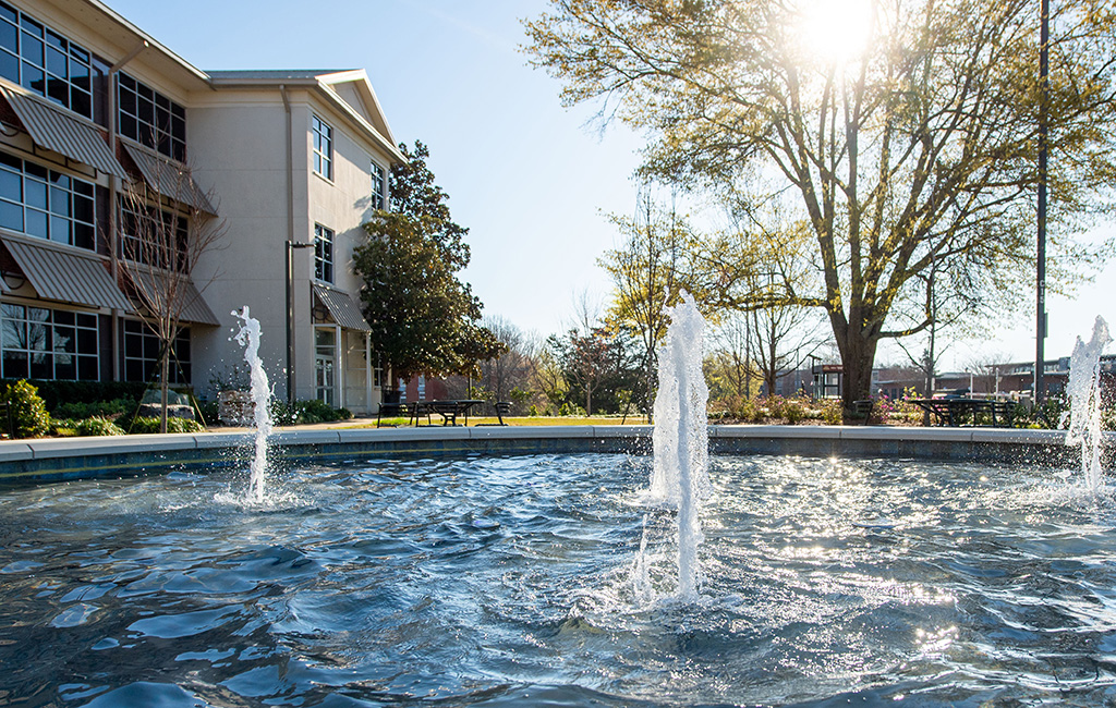 Water fountains in a circular pool outside a building, with sunlight shining through trees and benches visible in the background.