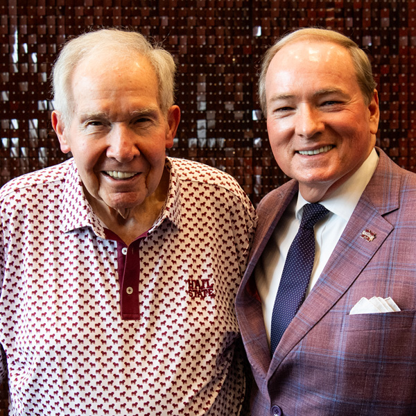 Two men stand side by side, smiling at the camera. The man on the left wears a patterned polo shirt, and the man on the right wears a suit and tie with a pocket square.