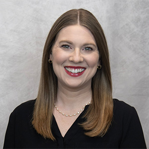 A woman with straight, shoulder-length brown hair, wearing a black top, a pearl necklace, and small hoop earrings, smiles in front of a light gray background.
