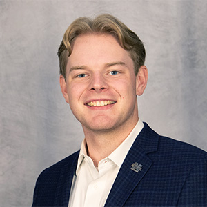 A man with light hair, wearing a navy blazer and white shirt, smiles in front of a gray textured backdrop.