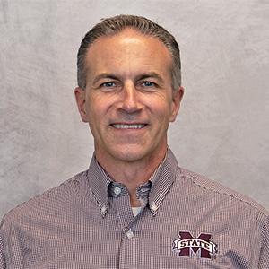 Middle-aged man smiling at the camera, wearing a maroon checkered shirt with a Mississippi State University logo, standing in front of a plain gray background.