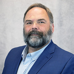 A man with a beard and mustache wearing a blue checked suit jacket and light blue shirt poses in front of a light gray background.
