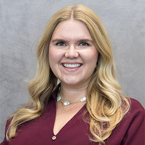 A woman with long blonde hair, wearing a maroon blouse and a pearl necklace, smiles in front of a gray background.