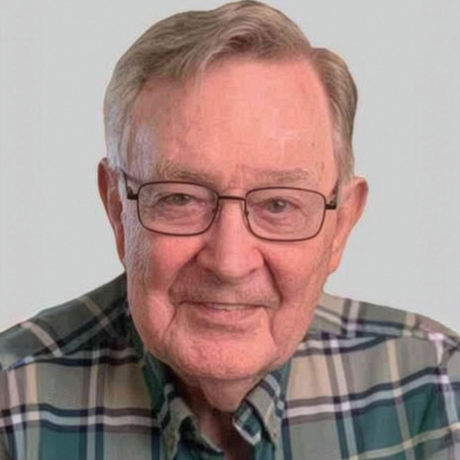 An elderly man with glasses and gray hair smiles at the camera, wearing a green and white plaid shirt against a plain light background.