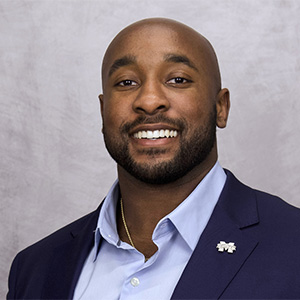 A man in a navy blazer and light blue shirt smiles at the camera against a light gray background.