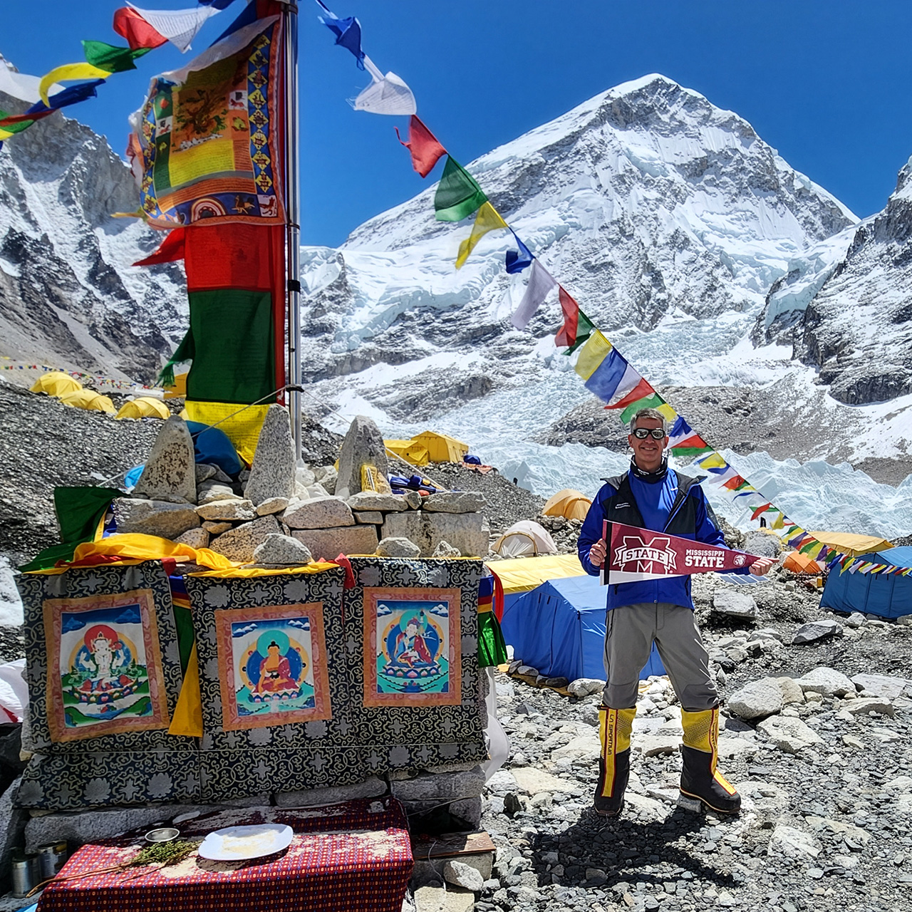 A person stands at Everest Base Camp holding a Mississippi State banner, surrounded by prayer flags, colorful tents, and Himalayan mountains in the background.