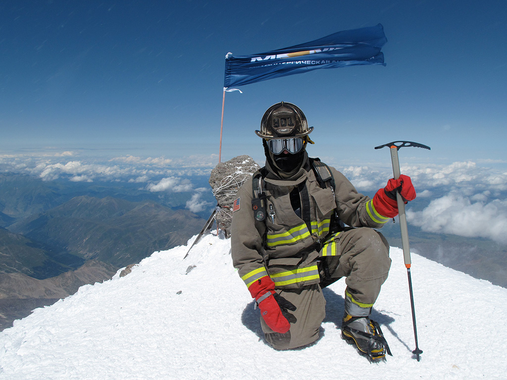A person in full firefighter gear kneels on a snowy mountain summit, holding an ice axe, with a blue flag and mountains visible in the background.