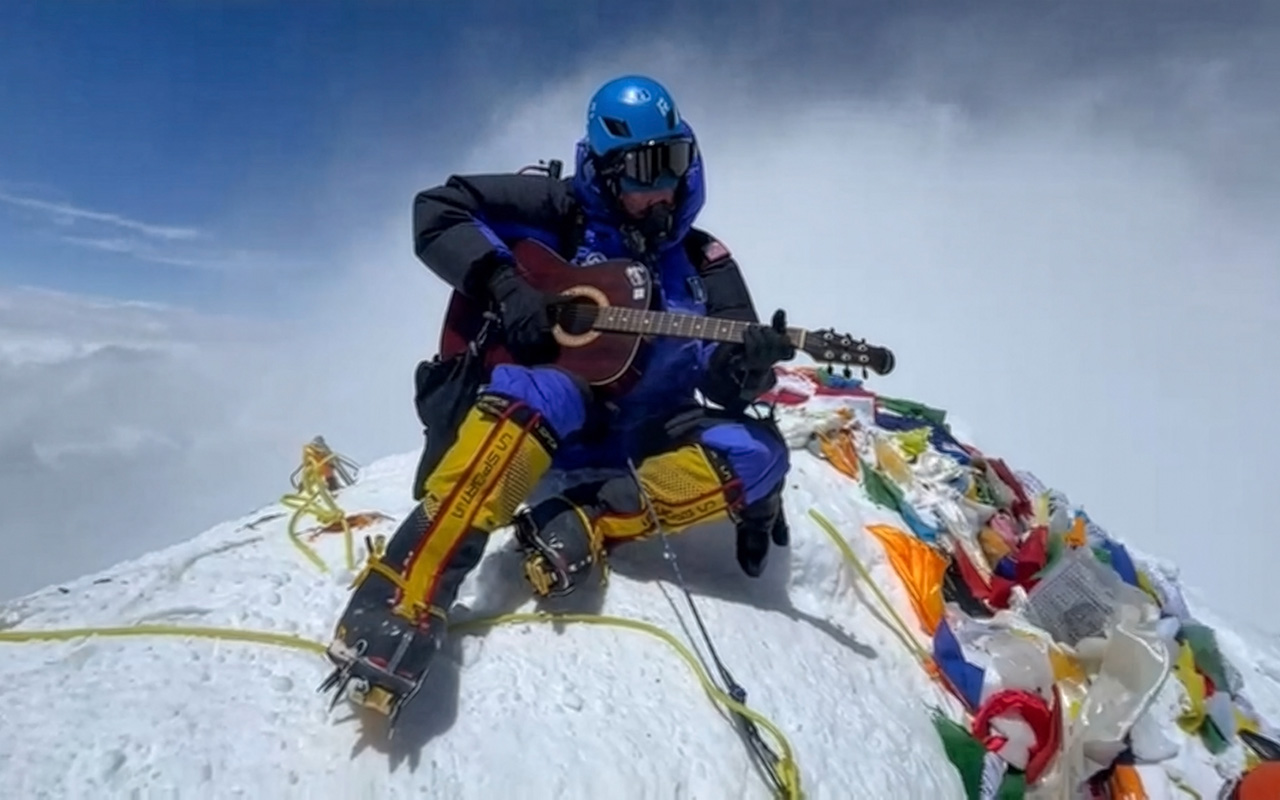 A climber in full gear plays a guitar while sitting on a snowy mountain summit surrounded by colorful prayer flags.