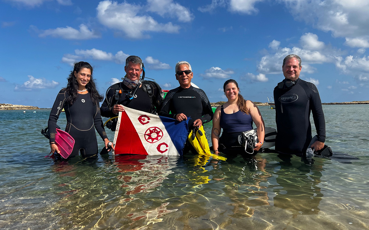 Five people in wetsuits stand in shallow water, holding scuba gear and an Explorers Club flag, with a blue sky and clouds in the background.