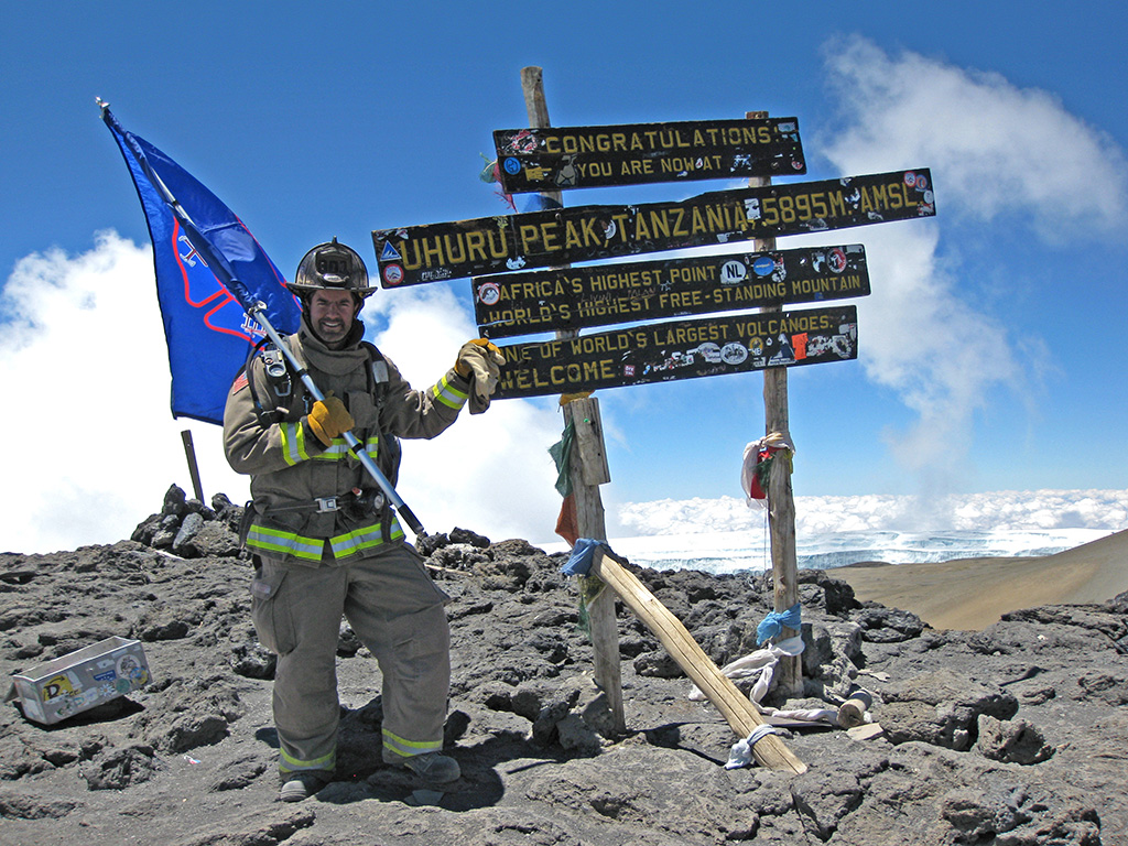 A person in firefighter gear holds a blue flag and poses next to the sign marking the summit of Uhuru Peak, Mount Kilimanjaro, with clouds and blue sky in the background.