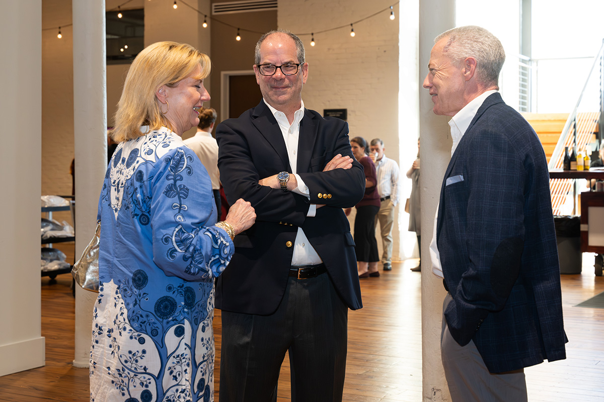 Three people in business attire stand indoors having a conversation at a social event, with other attendees visible in the background.