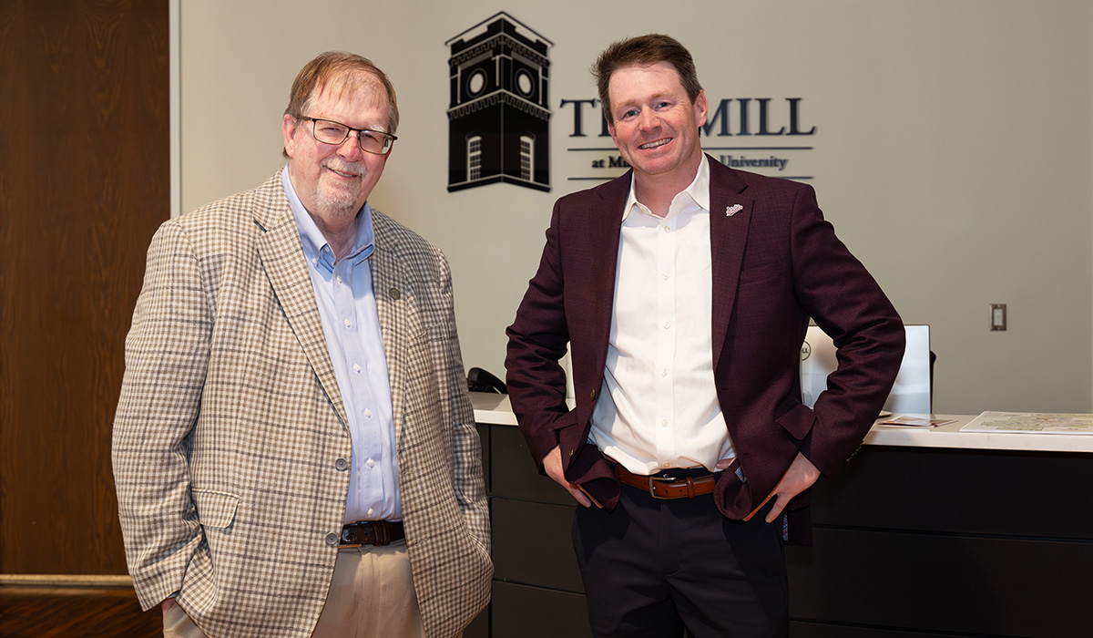 Two men in business attire stand and smile inside an office lobby with a sign reading "The Mill" visible on the wall behind them.