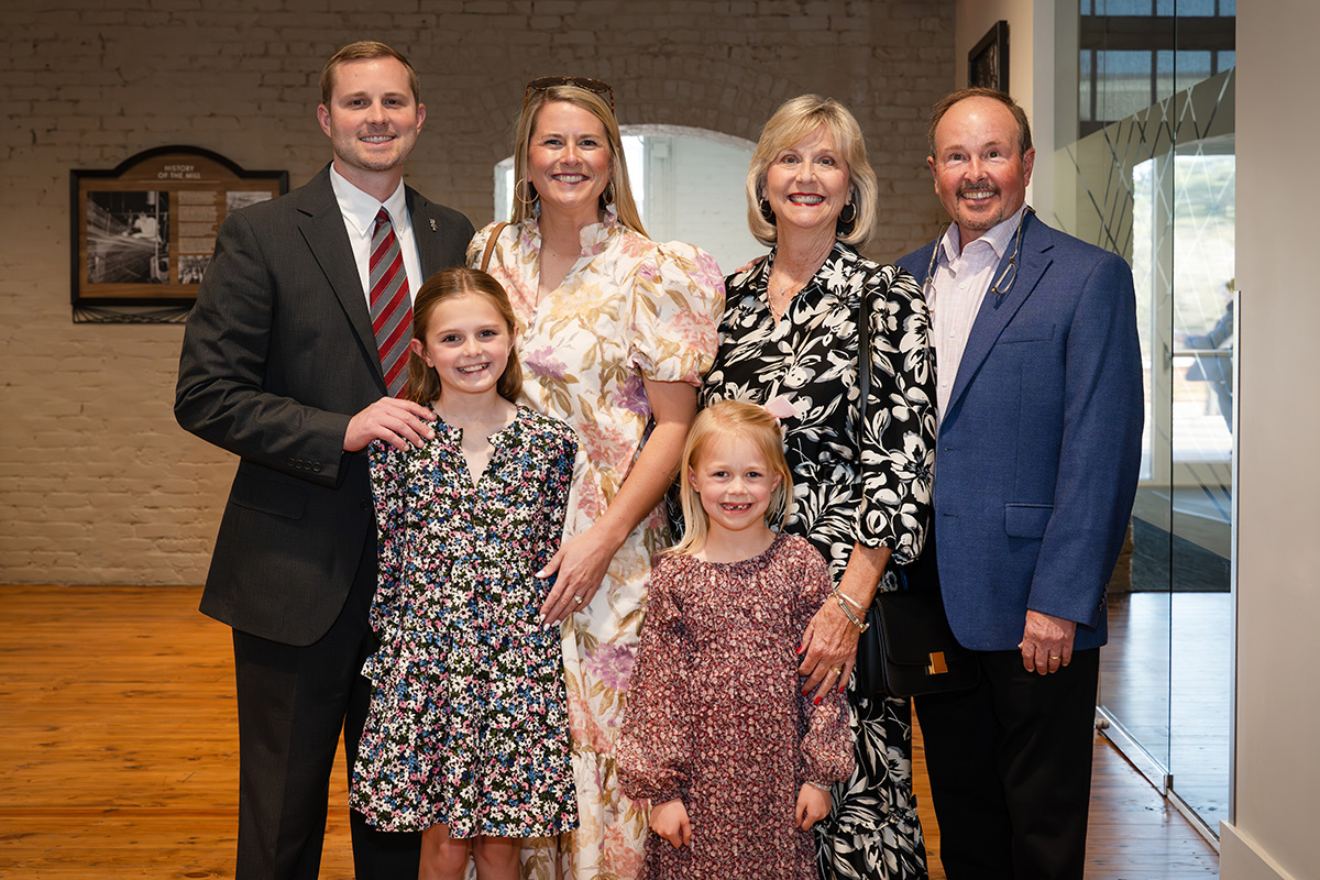 A group of three adults and two children pose indoors, dressed in formal and semi-formal attire, standing on a wooden floor with a brick wall and window in the background.