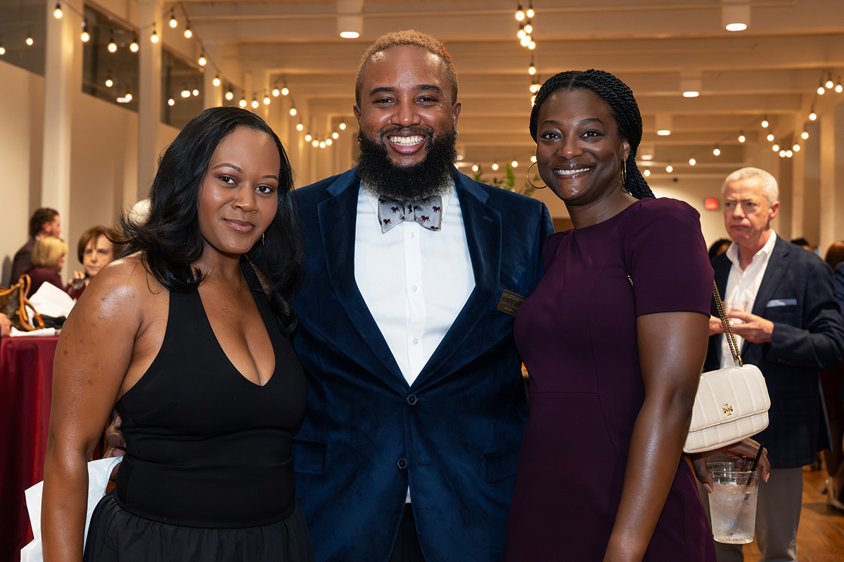 Three people pose together and smile at an indoor event with string lights; other guests can be seen in the background.
