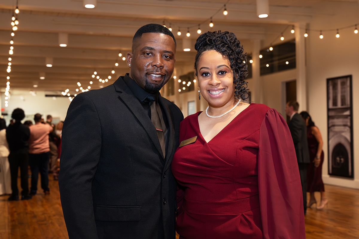 A man in a black suit and a woman in a burgundy dress pose and smile at an indoor event with string lights and guests in the background.