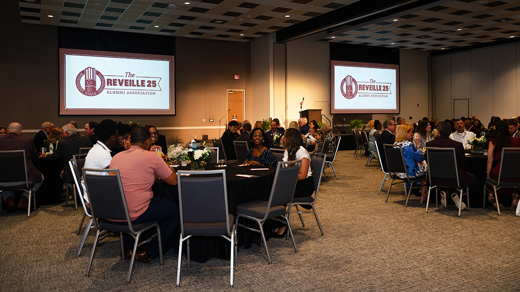 People seated at round tables in a banquet hall for the Reveille 25 Alumni Association event, with presentation screens displaying the event logo.