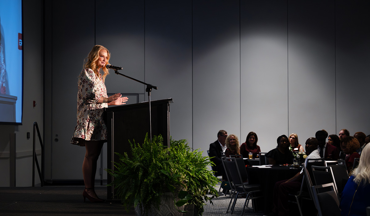 A woman speaks at a podium on stage during an event, with an audience seated at round tables and a fern in front of the podium.