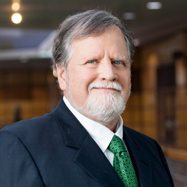 Older man with gray hair and beard, wearing a dark suit, white shirt, and green patterned tie, standing indoors with a blurred background.