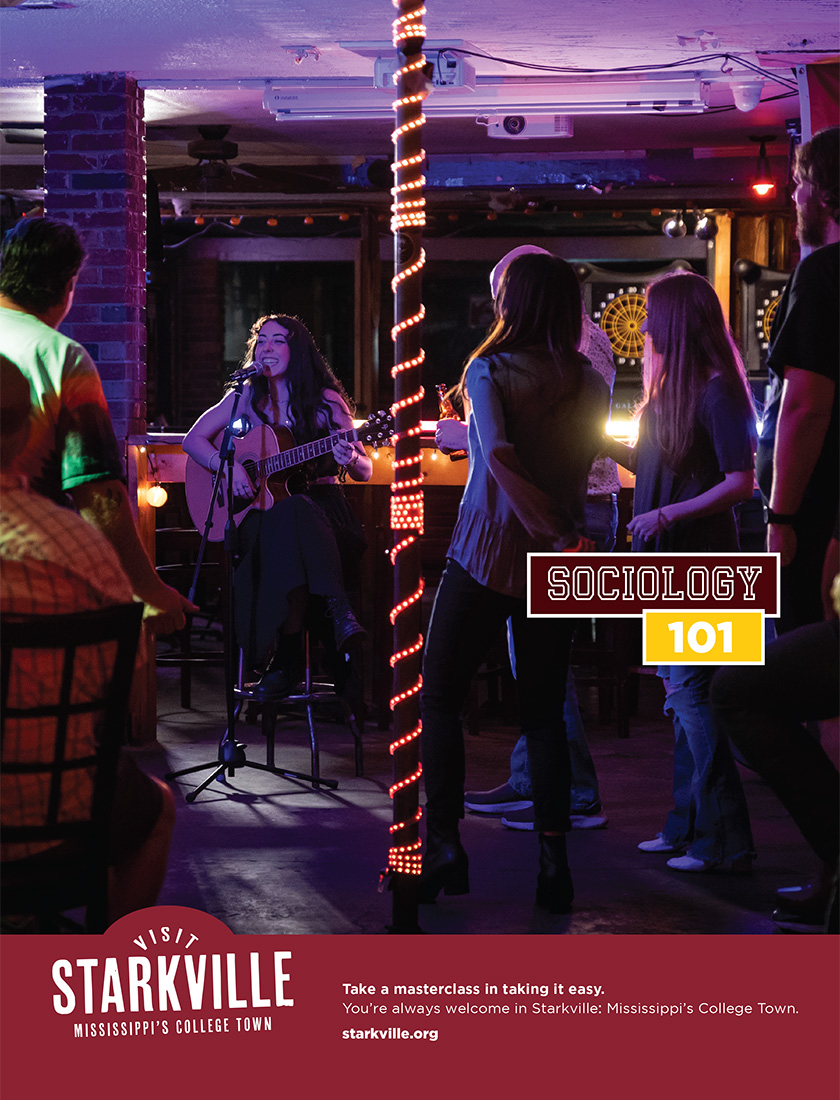 A woman plays guitar and sings on stage in a dimly lit bar while an audience watches. Text on the image promotes sociology and visiting Starkville, Mississippi's college town.