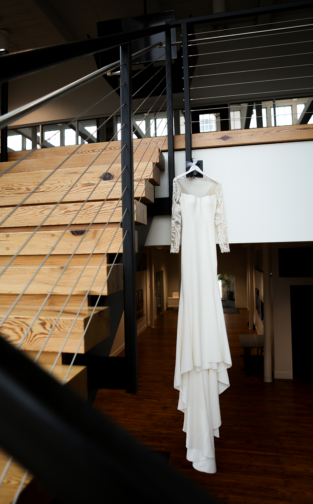 A white, long-sleeved wedding dress with lace detail hangs on a hanger from a staircase in a modern interior space.