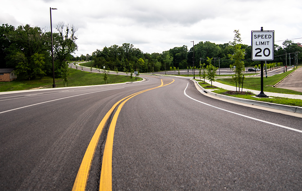 Curved, freshly paved road with a double yellow line and a posted speed limit sign of 20 mph; trees and grass line both sides under an overcast sky.