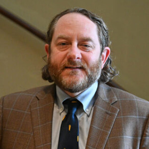 A man with wavy brown hair and a beard is wearing a brown plaid blazer, light blue shirt, and patterned tie, standing indoors in front of a plain background.