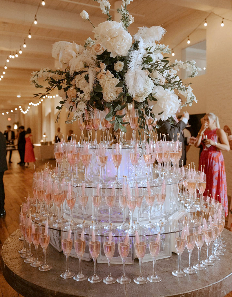 A tiered display of filled champagne glasses surrounds a large floral arrangement in a decorated indoor event space with guests in the background.