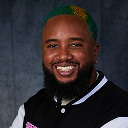 A man with a beard and green and yellow dyed hair smiles, wearing a black and white jacket against a dark background.