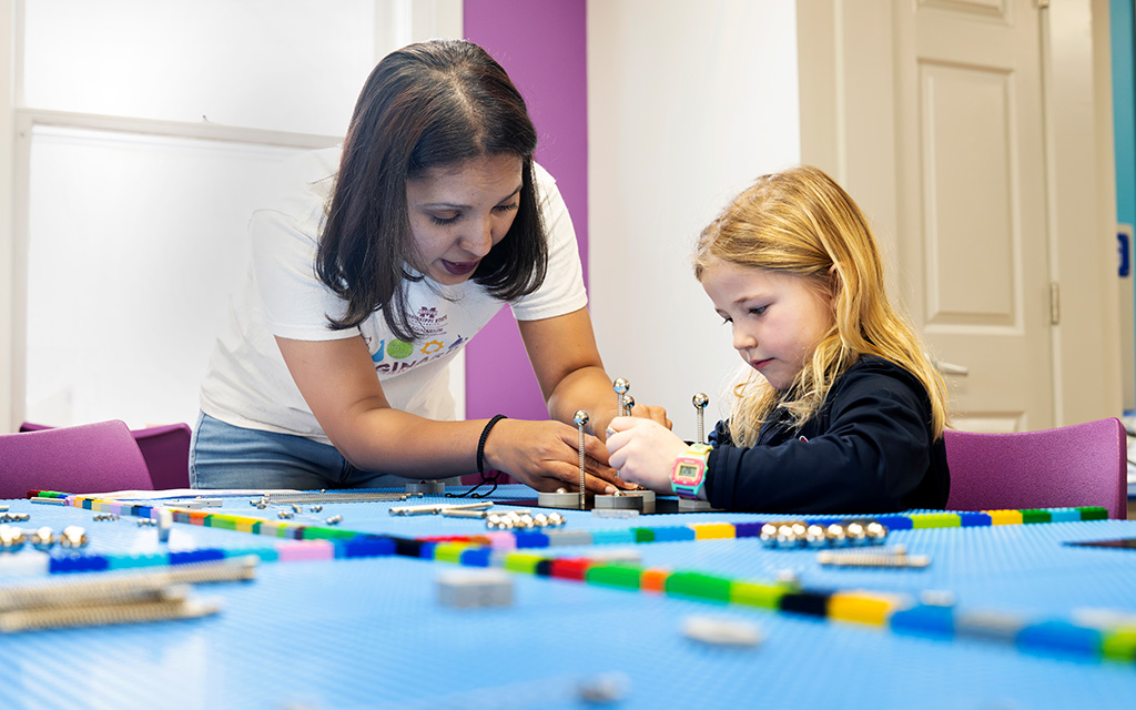 An adult helps a young girl assemble a small robotic project on a blue table covered with LEGO pieces and robotics components in a classroom.