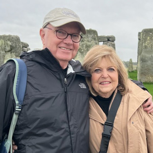 An older man and woman stand close together, smiling in front of large stone structures at Stonehenge on a cloudy day.