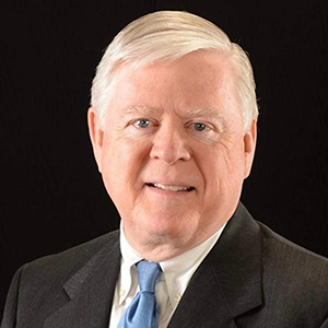 An older man with short gray hair wearing a dark suit, white shirt, and light blue tie poses against a plain black background.