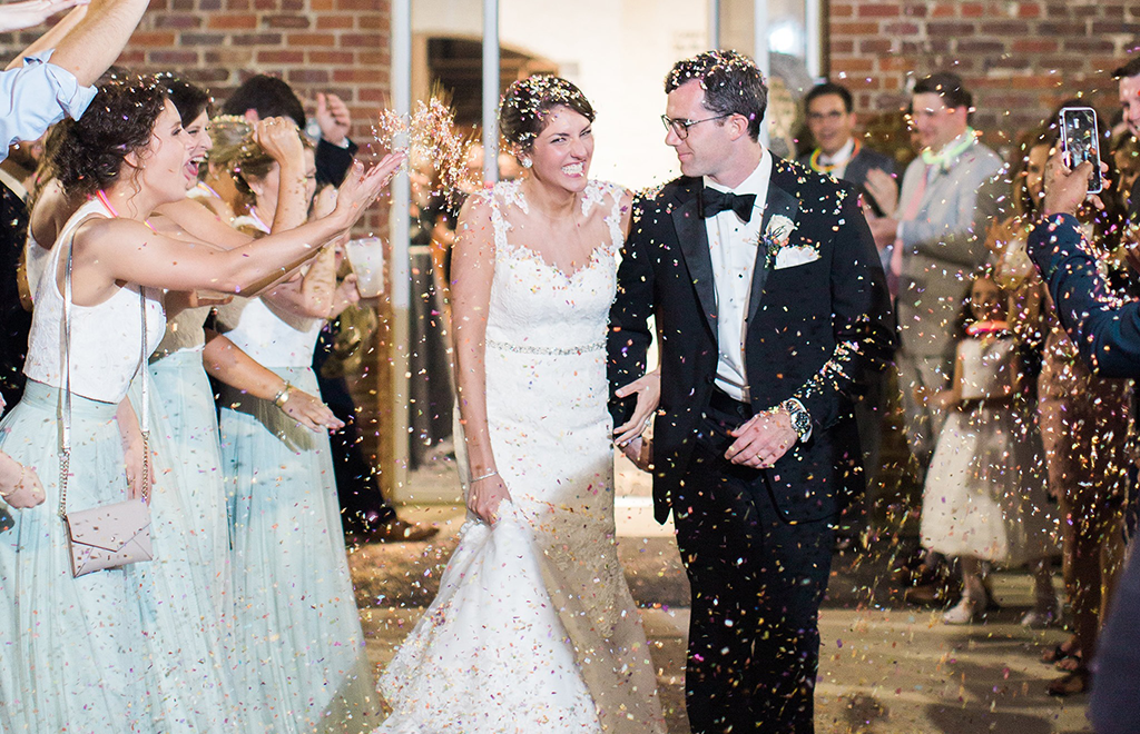 A bride and groom walk hand in hand while guests throw confetti at them during a wedding celebration.