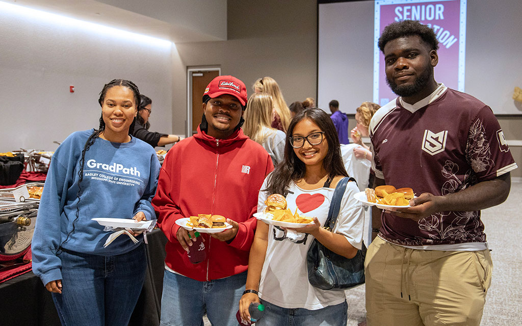 Four people are standing indoors, smiling at the camera and holding plates of food at a casual event.