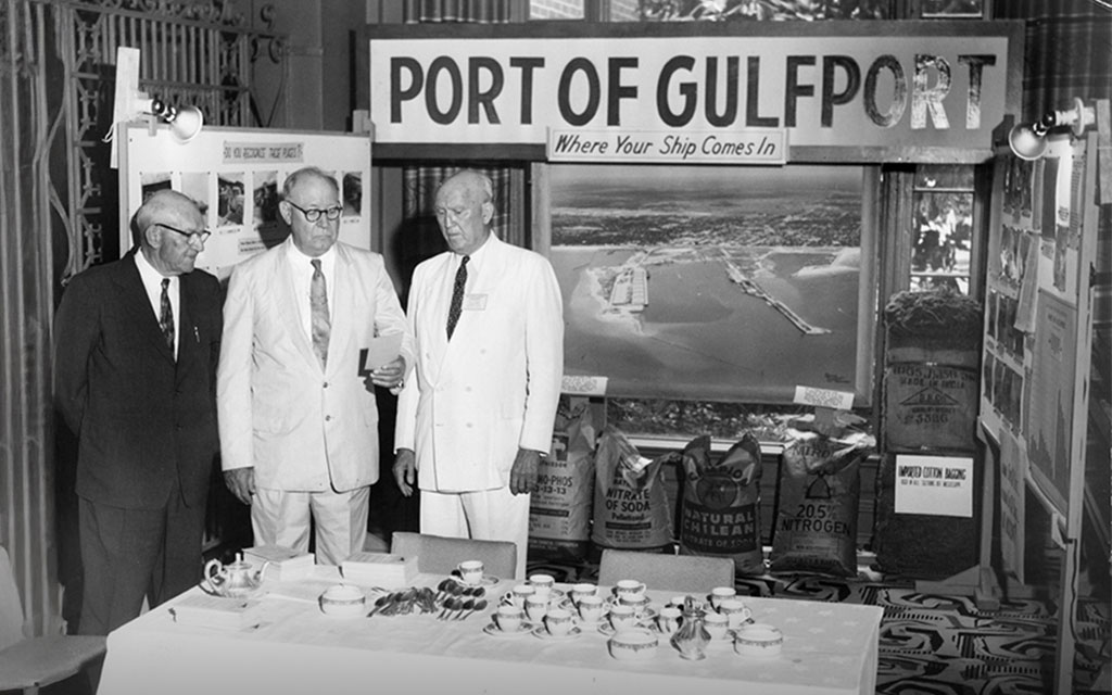 Three men in suits stand at a display booth for the Port of Gulfport, featuring an aerial photo, informational materials, and bags of fertilizer.