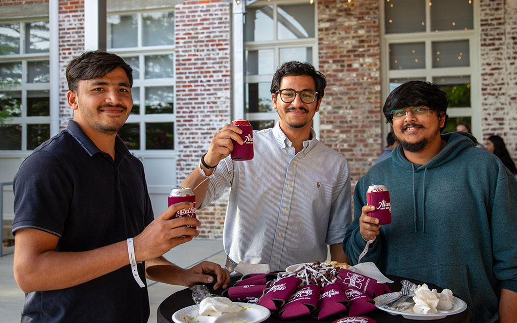 Three men stand around a tall table outdoors, holding canned drinks with maroon koozies, smiling at the camera. Used plates and extra koozies are on the table.
