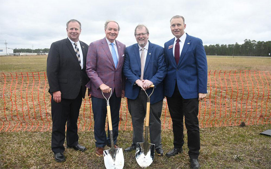 Four men in suits stand on grass behind an orange barrier, two holding shovels, at a groundbreaking ceremony on a cloudy day.