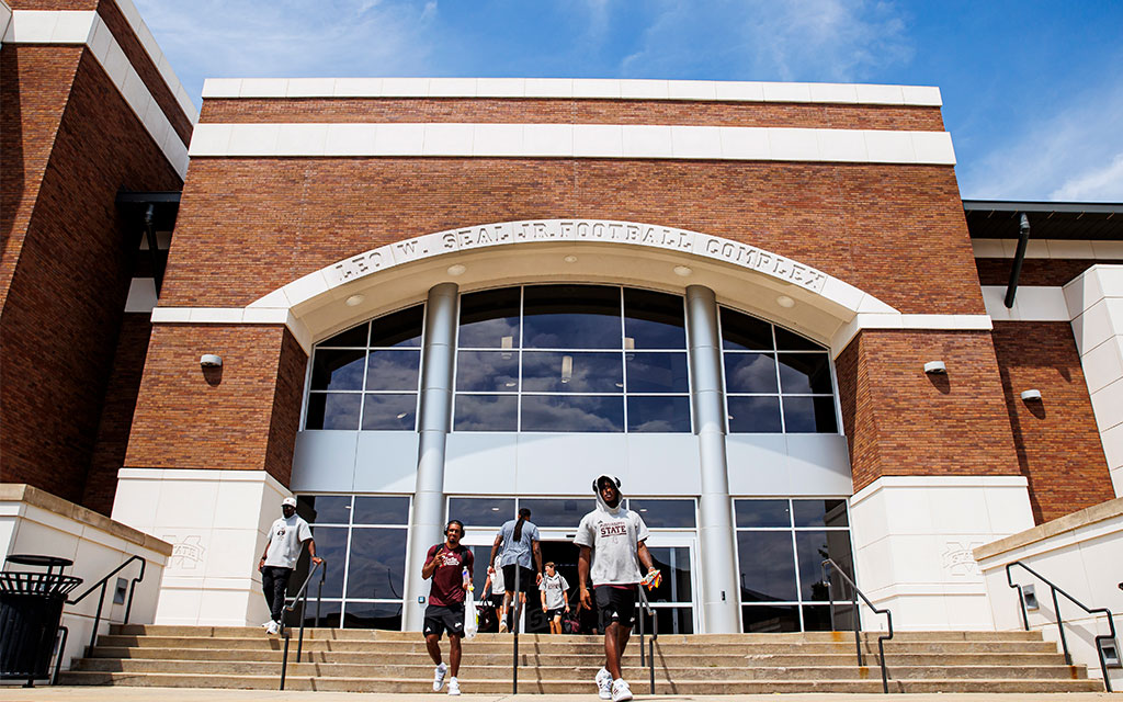Several people walk down steps outside a large brick building labeled “Leo W. Seal Jr. Football Complex” under a blue sky.