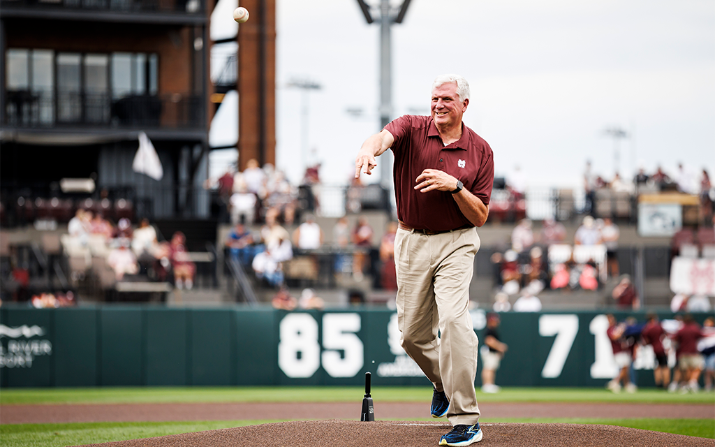 An older man throws a ceremonial first pitch on a baseball field, wearing a maroon polo and khaki pants, with spectators in the background.