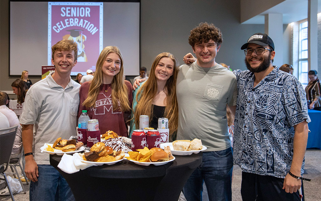 Five young adults stand smiling behind a table with food and drinks at a “Senior Celebration” event, with a projector screen and other attendees visible in the background.