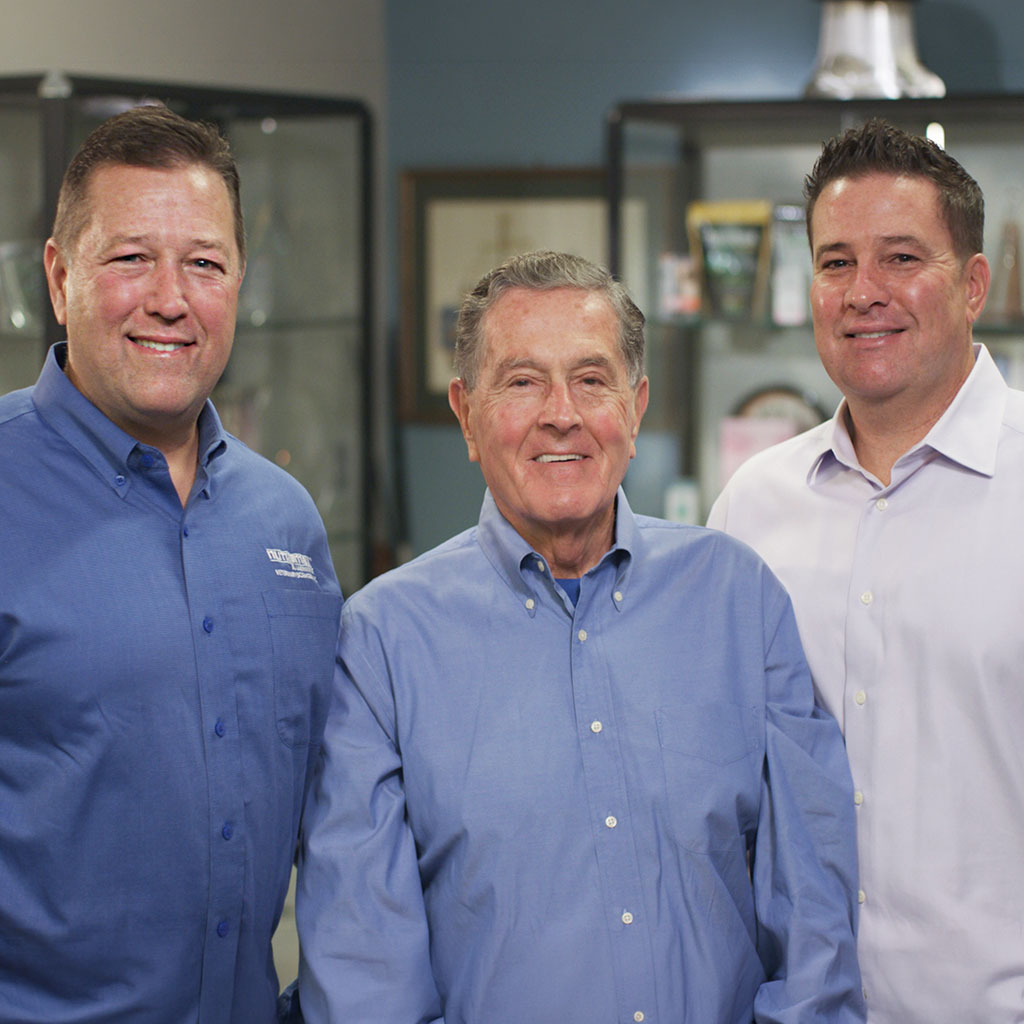 Three men stand indoors, all wearing blue button-down shirts, posing and smiling for the camera with display cabinets visible in the background.