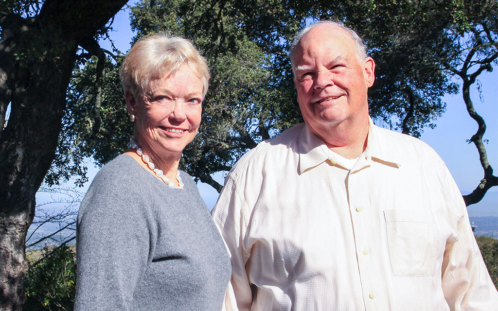 An older woman and man stand outdoors in sunlight, smiling at the camera with trees and blue sky in the background.