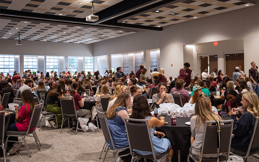 Large group of people seated at round tables in a spacious, well-lit event hall, engaged in conversation during a social gathering or meal.